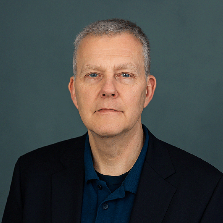 A man with salt and pepper hair, wearing a blue shirt and a dark jacket, sits in front of a green background